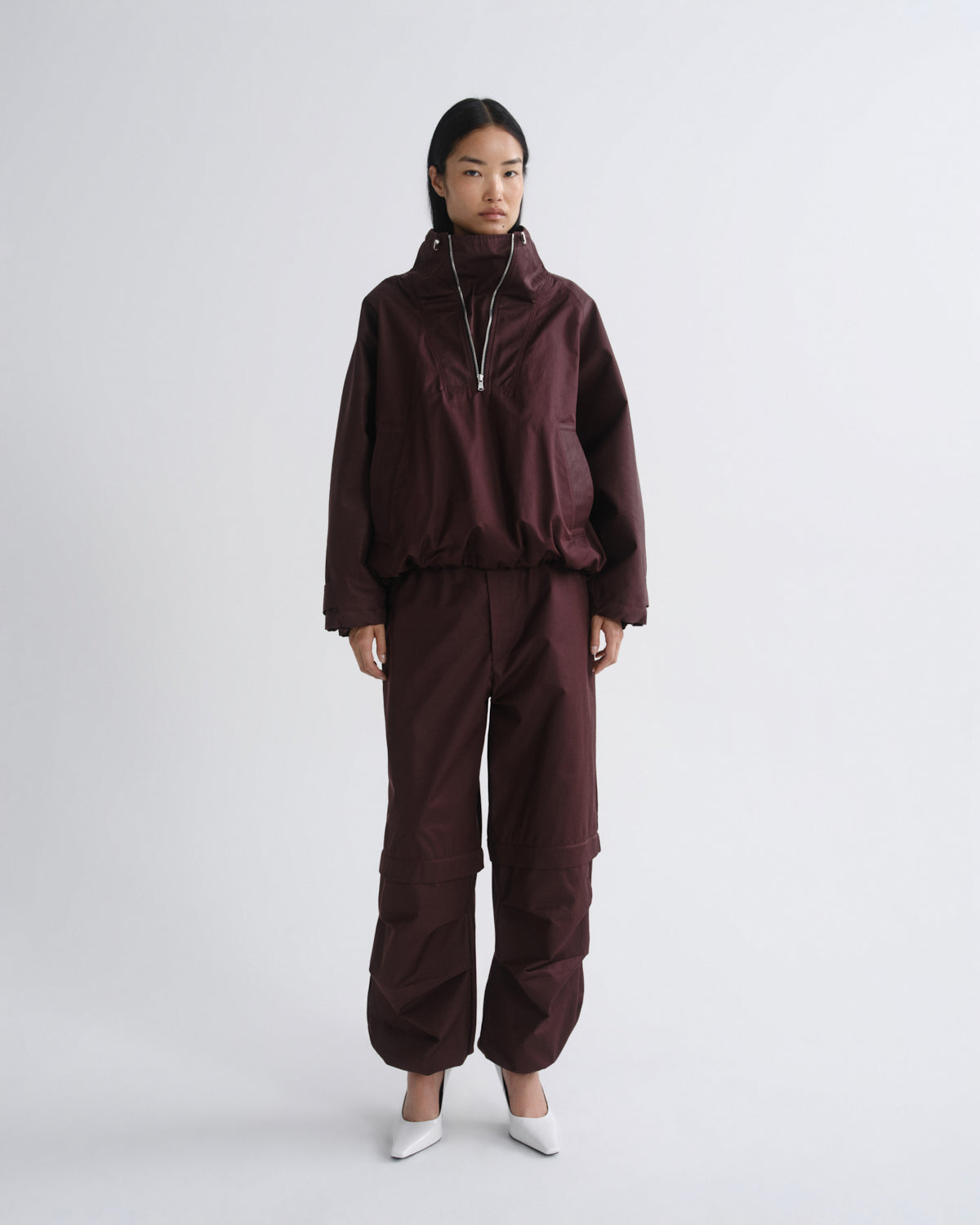 A model stands against a light gray studio background wearing a matching dark burgundy windbreaker jacket and cargo pants set, paired with white pointed shoes.