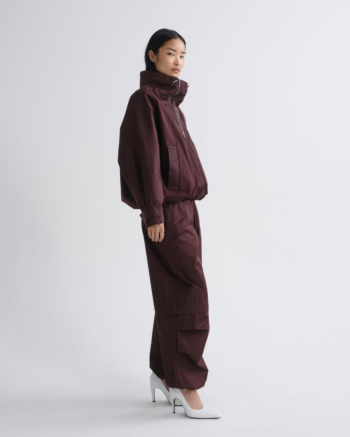 A model is shown in profile wearing a matching dark burgundy two-piece set consisting of a cropped zip-up jacket and wide-leg cargo pants, paired with white high-heeled shoes, against a light gray studio background.