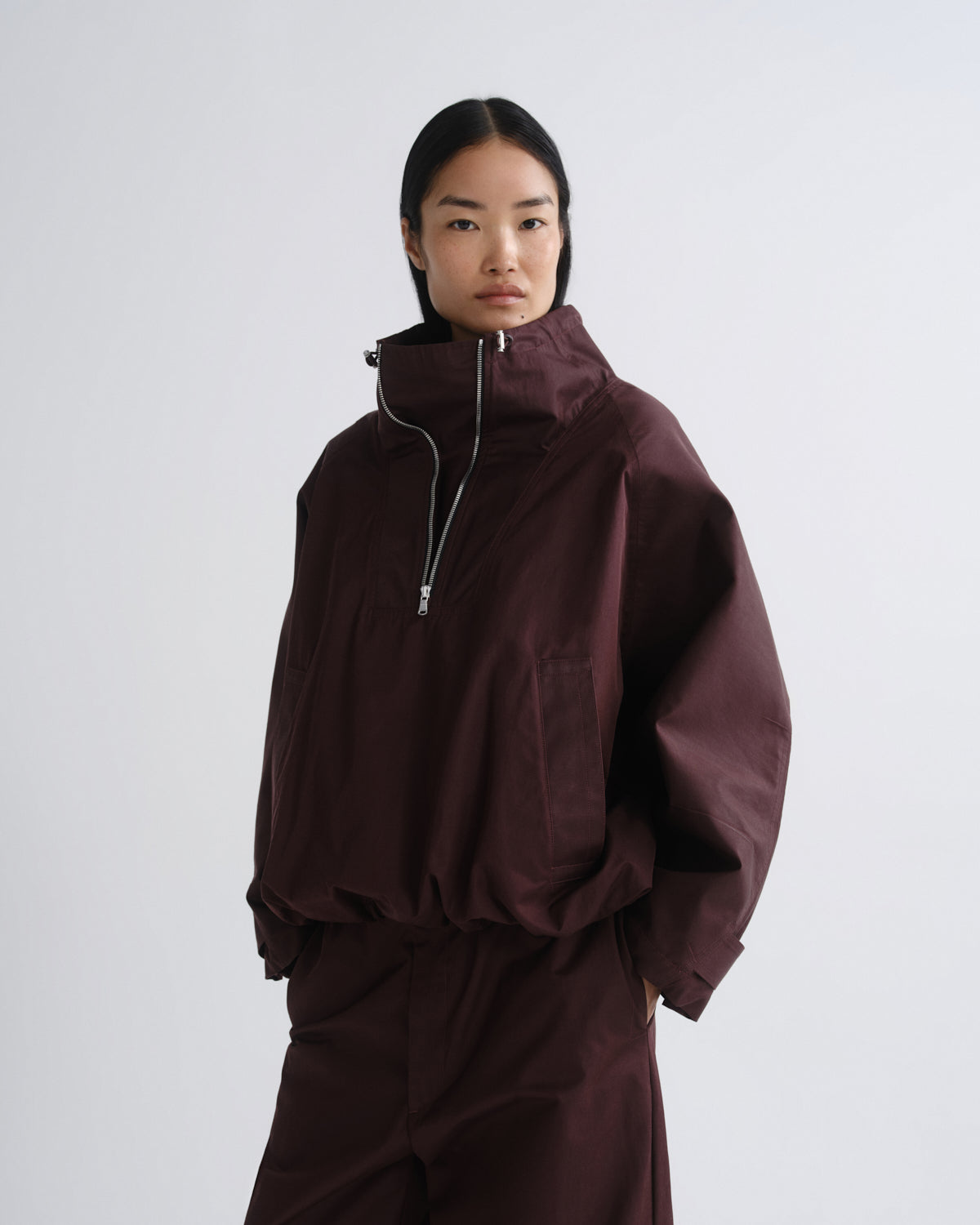 A model wearing a dark burgundy half-zip jacket with a high collar and matching dark pants, photographed against a neutral light gray studio background.