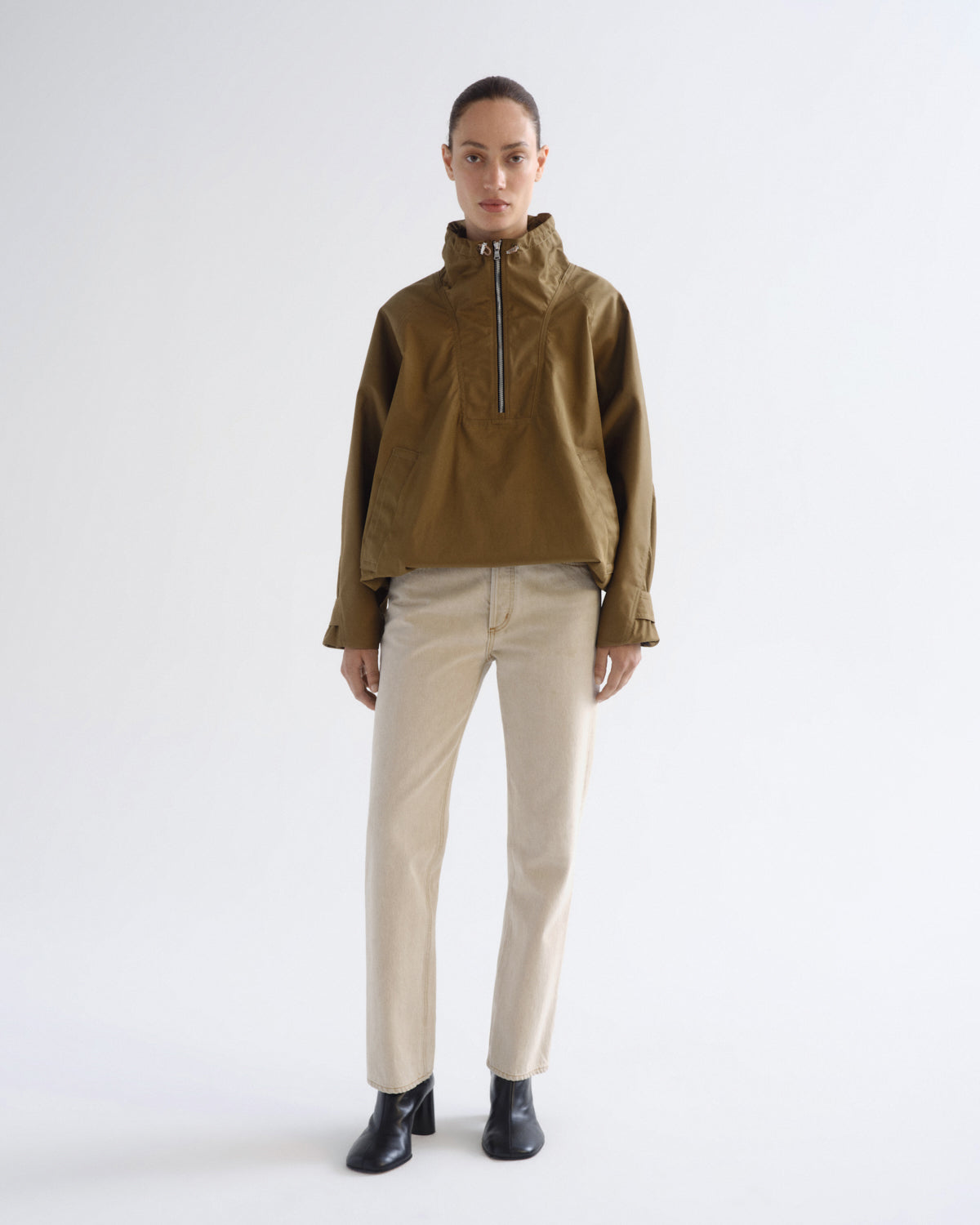 A model stands against a light gray studio background wearing a cropped olive-brown half-zip jacket, beige straight-leg pants, and black ankle boots.