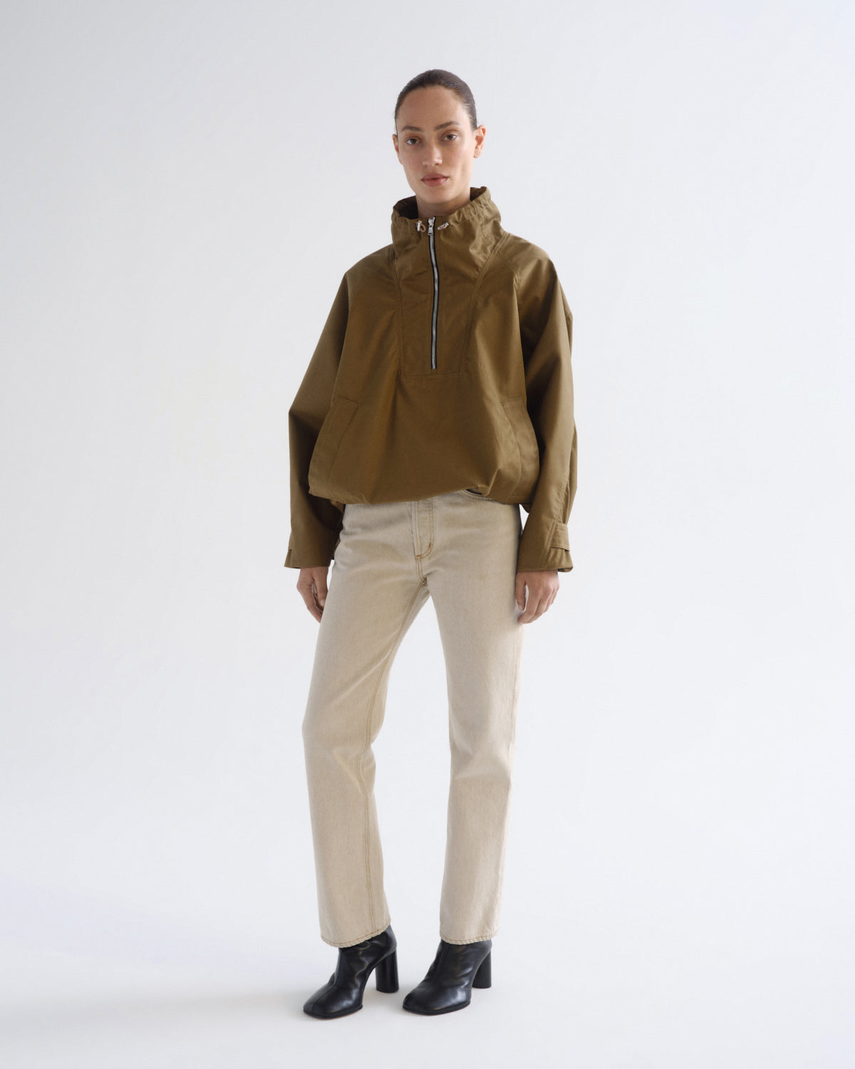 A model stands against a plain white studio background wearing a cropped olive-brown half-zip pullover jacket, light beige trousers, and black ankle boots.