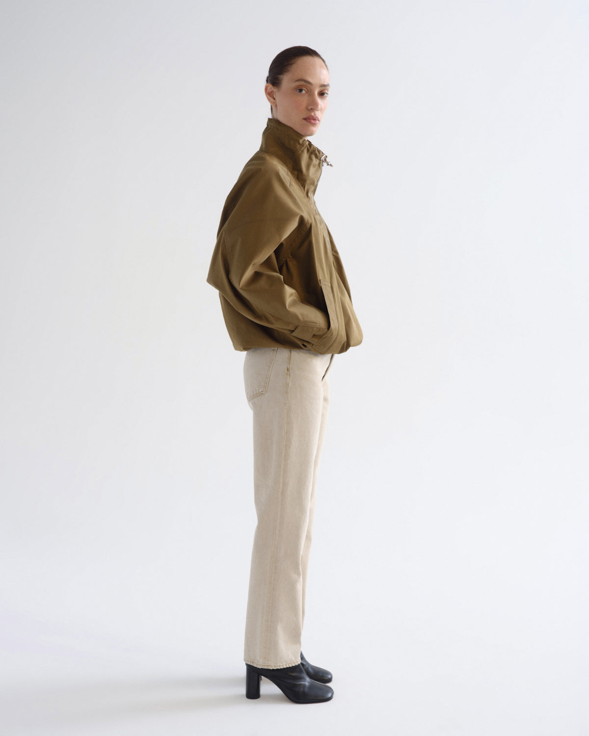 A model is shown in profile wearing a cropped olive green jacket, cream-colored trousers, and black high-heeled shoes against a plain white studio background.