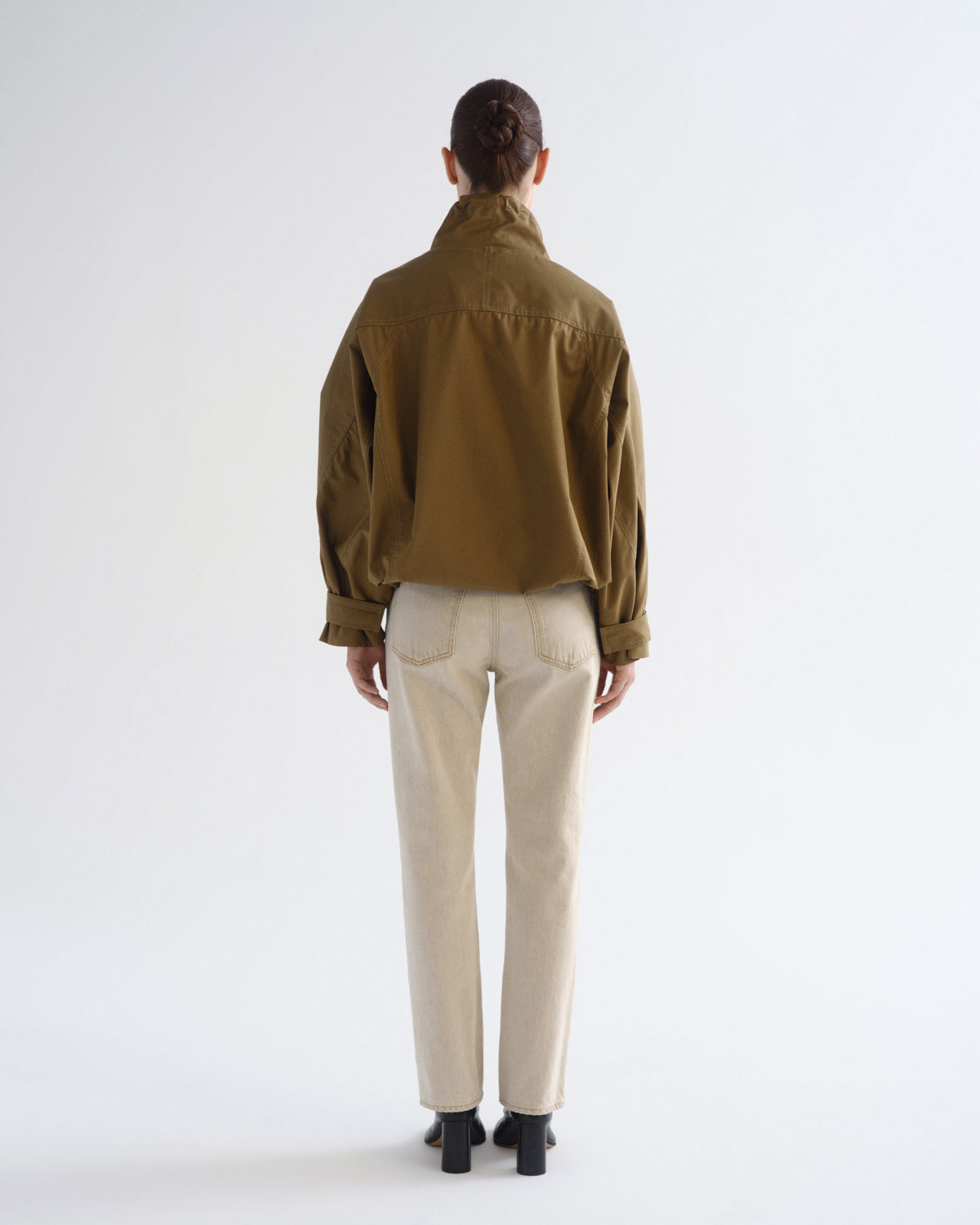 A model shown from the back wearing a cropped olive-brown jacket, cream-colored trousers, and black shoes, standing against a plain white studio background.