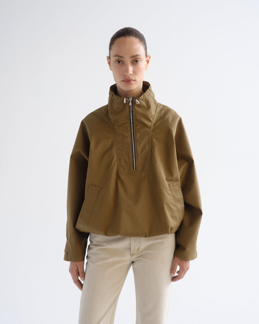 A model wearing a cropped olive-brown half-zip jacket with a high stand-up collar and beige trousers, standing against a plain light gray studio background.