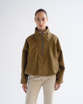 A model wearing a cropped olive-brown half-zip jacket with a high stand-up collar and beige trousers, standing against a plain light gray studio background.
