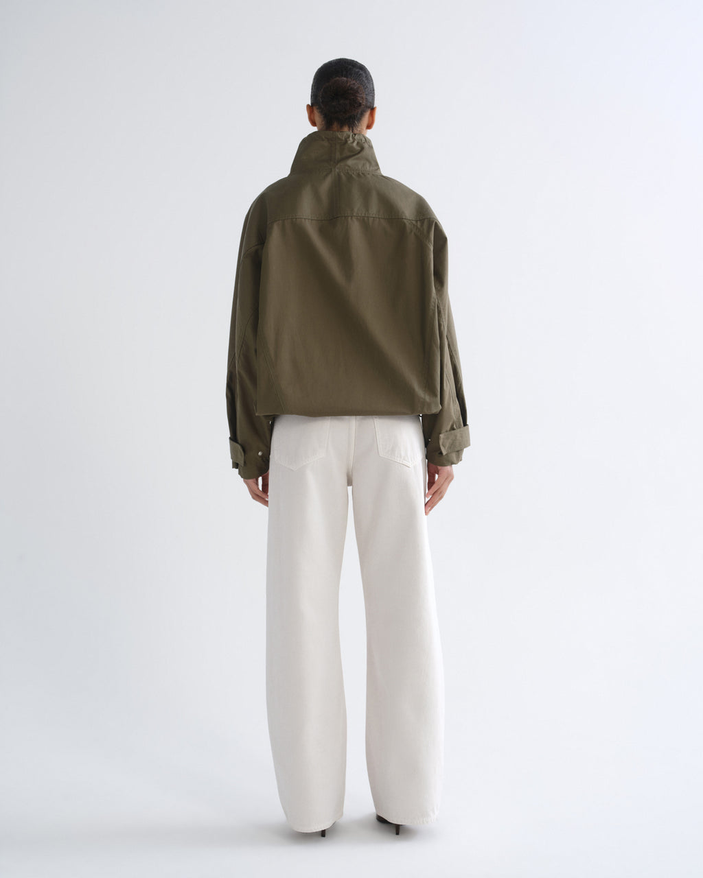 Person wearing olive green hooded pullover jacket and white pants, back view