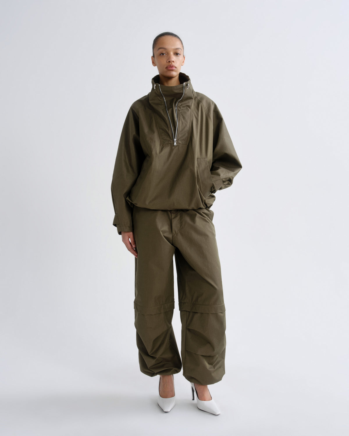 A model stands against a light gray studio background wearing an olive green two-piece tracksuit consisting of a high-collar zip-front jacket and matching wide-leg cargo pants, paired with white pointed heels.