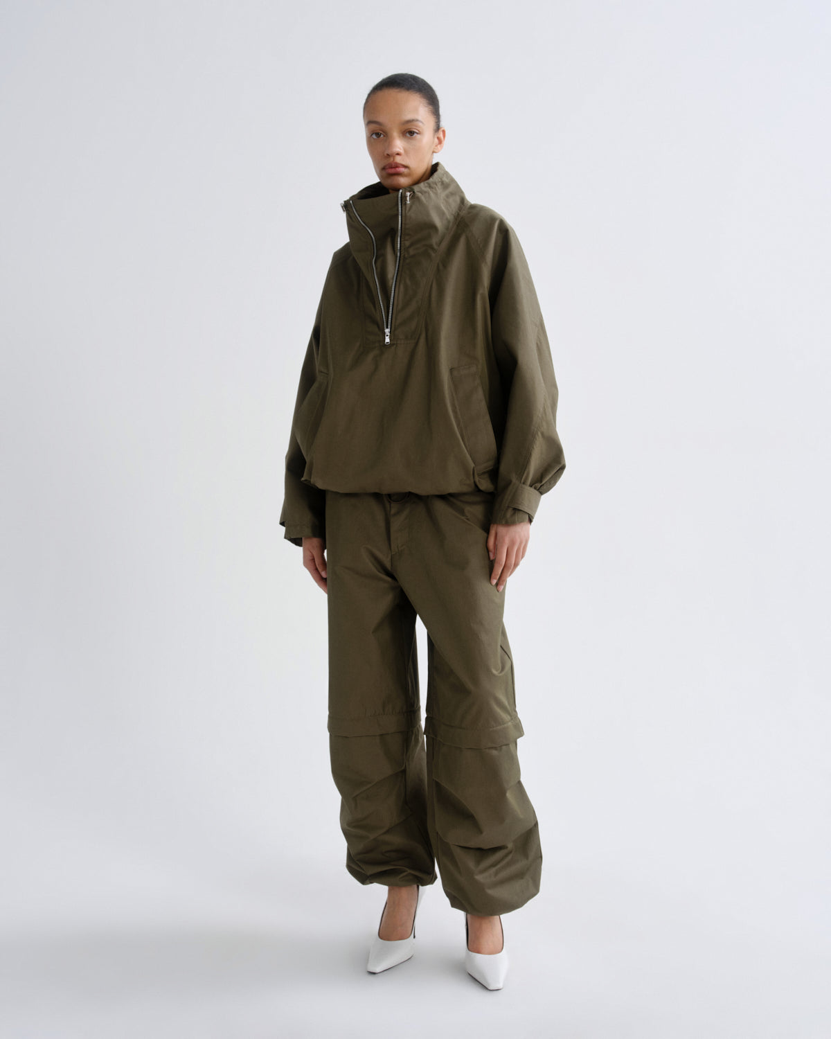 A model stands against a light gray studio background wearing an olive green hooded jacket with a half-zip front and matching wide-leg cargo pants, paired with white pointed shoes.