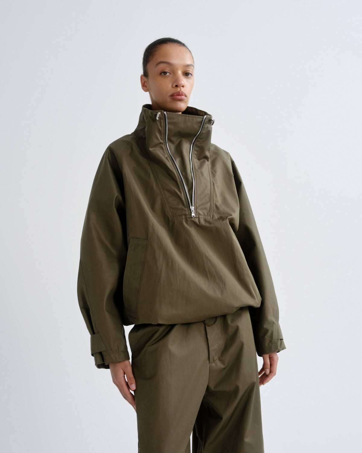 A model wearing an olive green lightweight anorak jacket with a high collar and half-zip, paired with matching olive green trousers, against a light gray studio background.