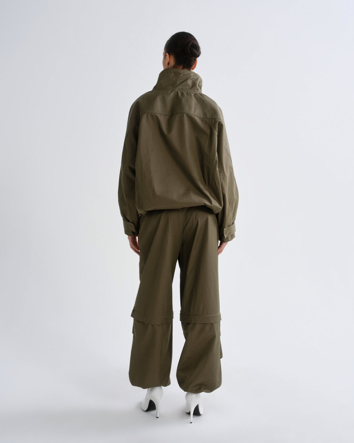 A model viewed from the back wearing an olive green utility jacket and matching wide-leg cargo pants, paired with white high-heeled shoes, against a plain light gray studio background.