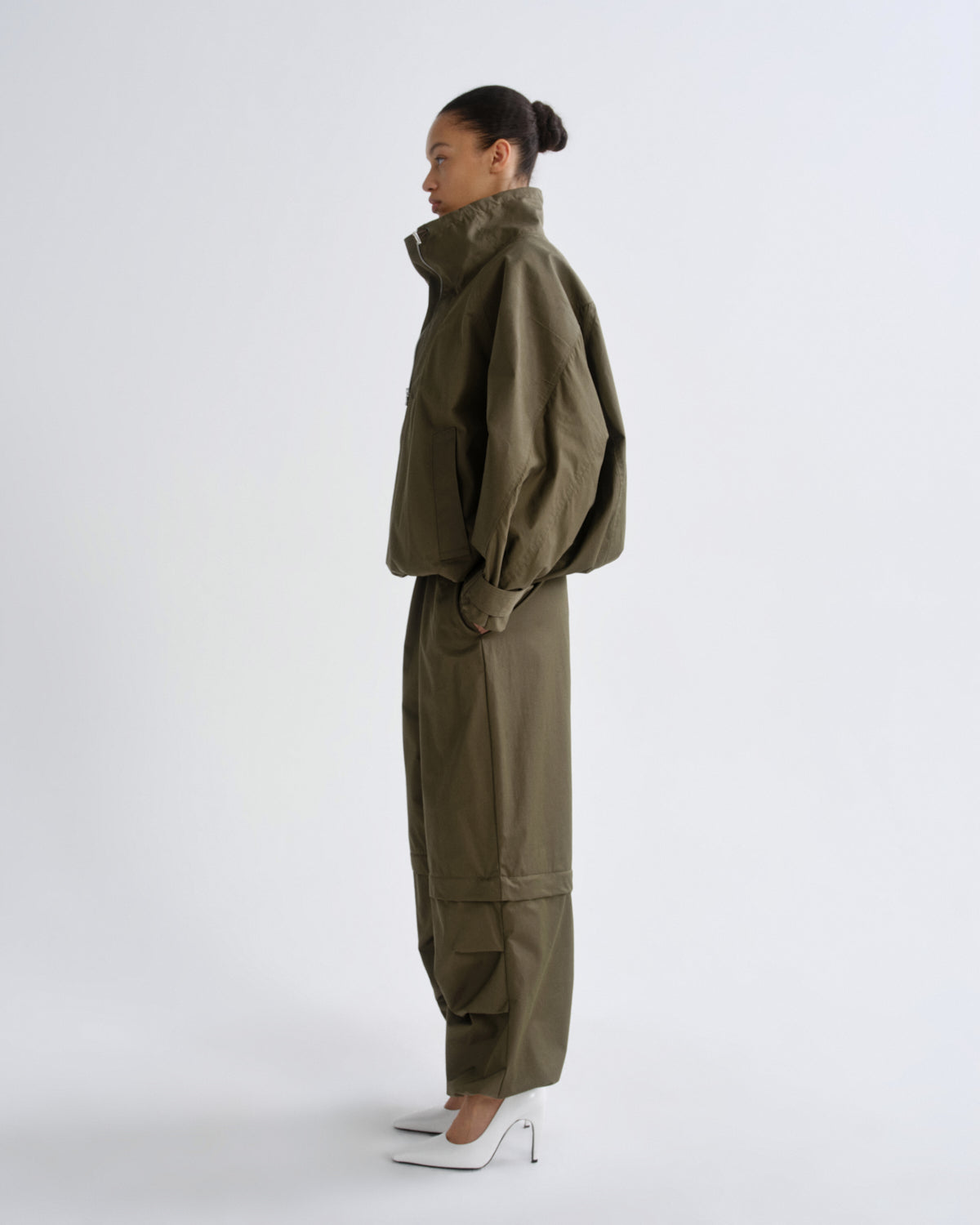 A model in profile wearing an olive green utility jacket and matching cargo pants, paired with white pointed-toe heels, against a plain light gray studio background.