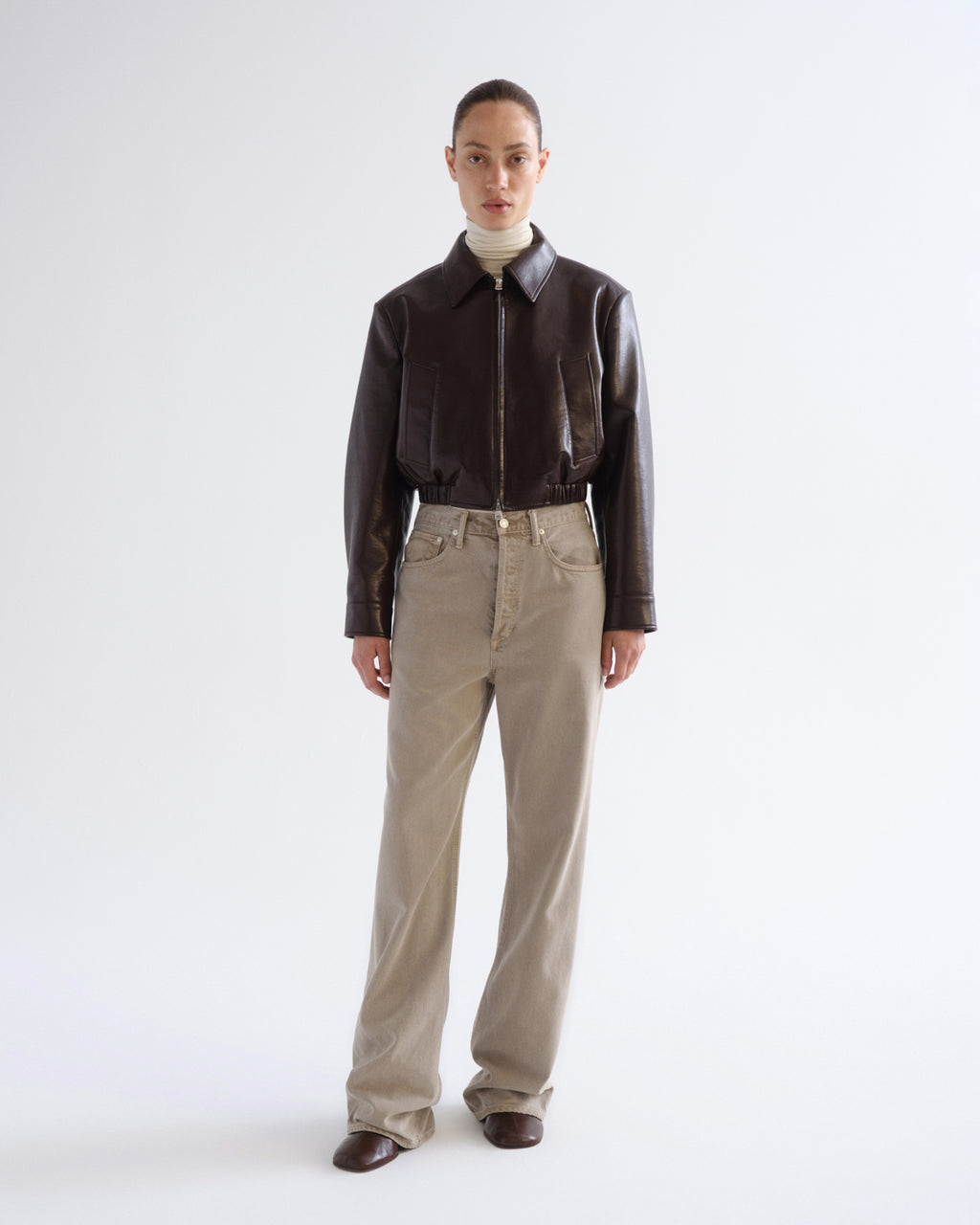 A model stands against a light gray studio background wearing a dark brown leather jacket, beige straight-leg jeans, and brown shoes.