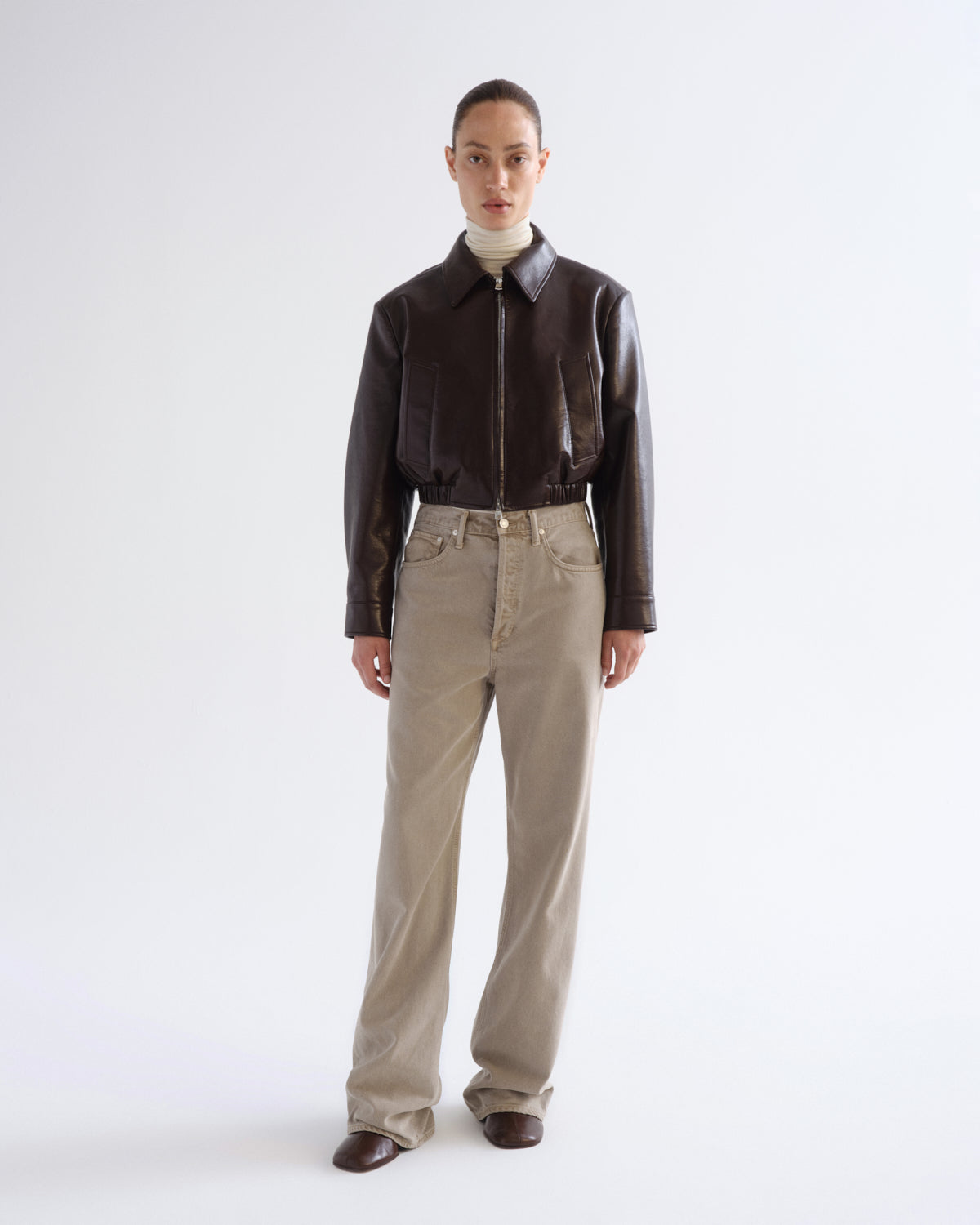 A model stands against a light gray studio background wearing a dark brown leather jacket, beige straight-leg jeans, and brown shoes.