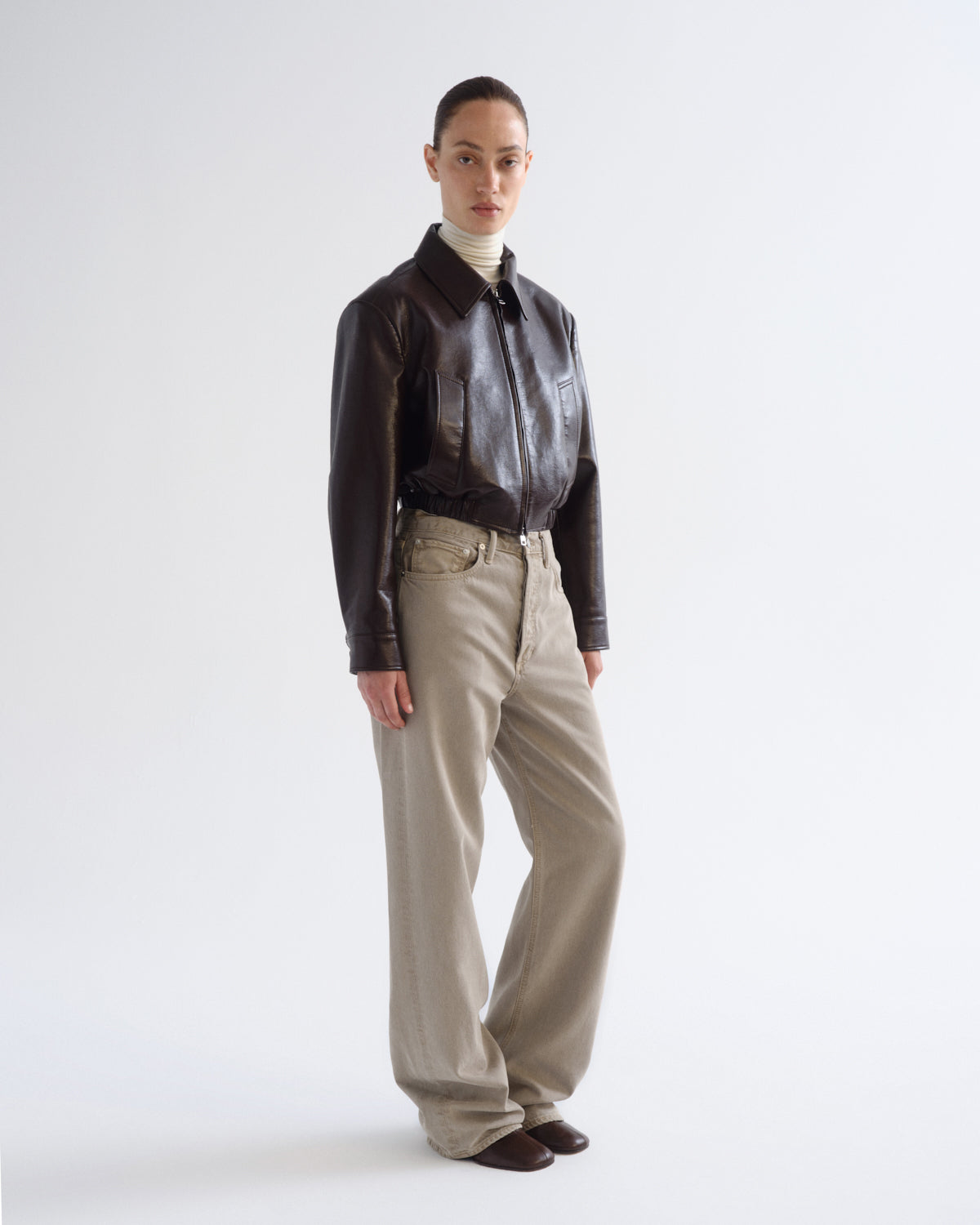 A model stands against a light gray studio background wearing a dark brown leather jacket, a cream ribbed turtleneck, beige wide-leg trousers, and dark brown shoes.