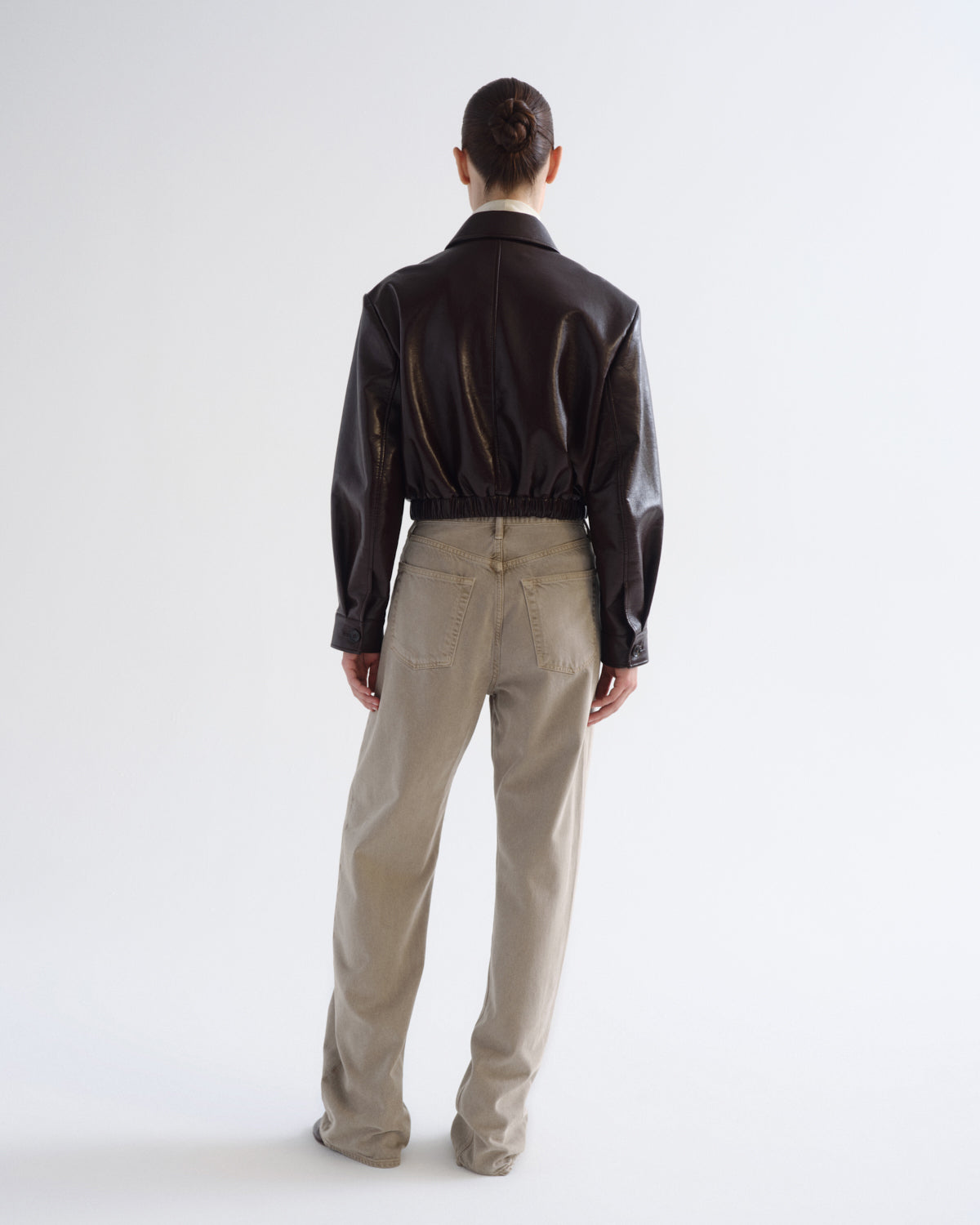 A model viewed from behind wearing a cropped black leather jacket and beige trousers, standing against a plain white studio background.