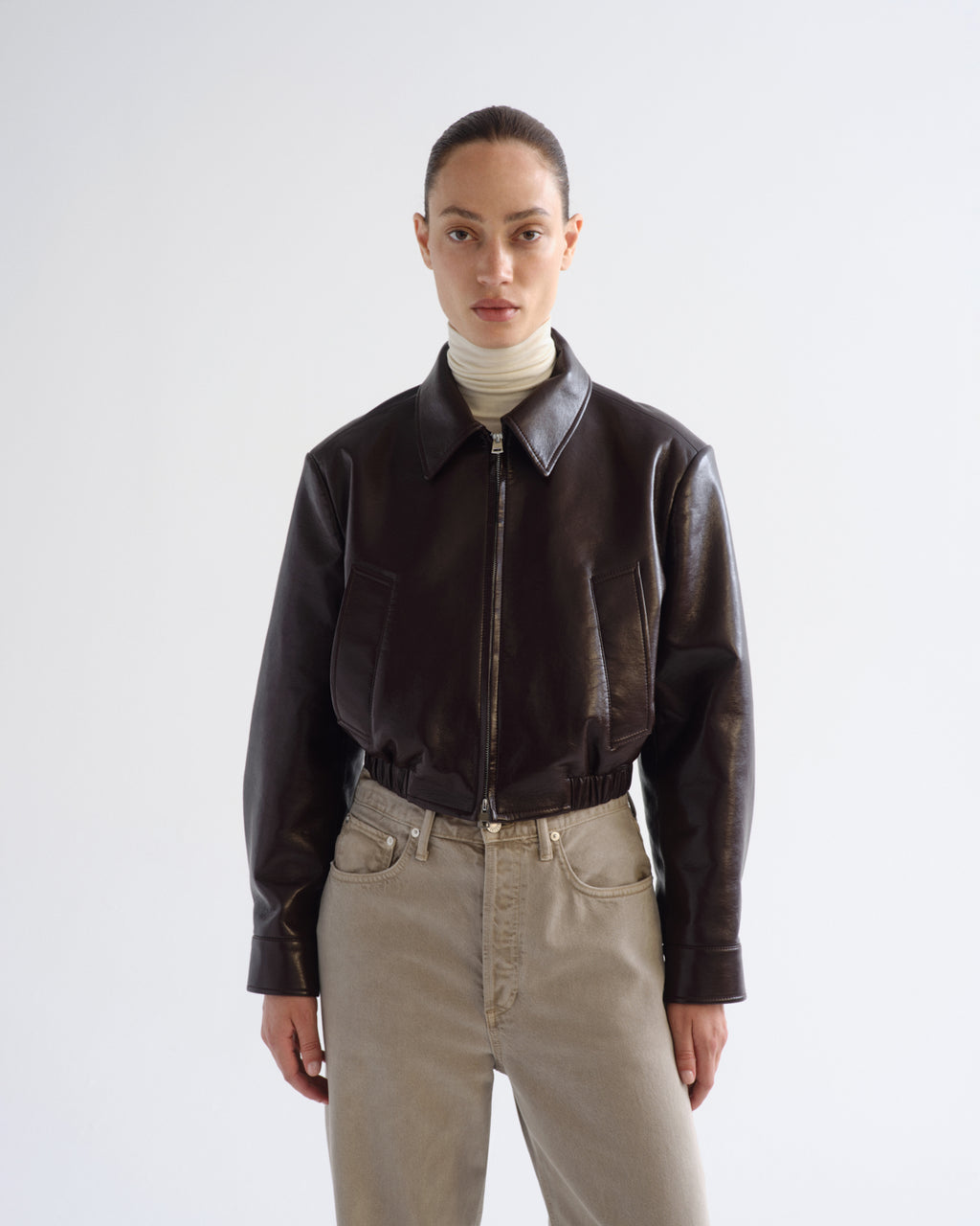A model wearing a dark brown leather cropped jacket with a front zipper, paired with a cream turtleneck and beige jeans, shot against a light gray studio background.