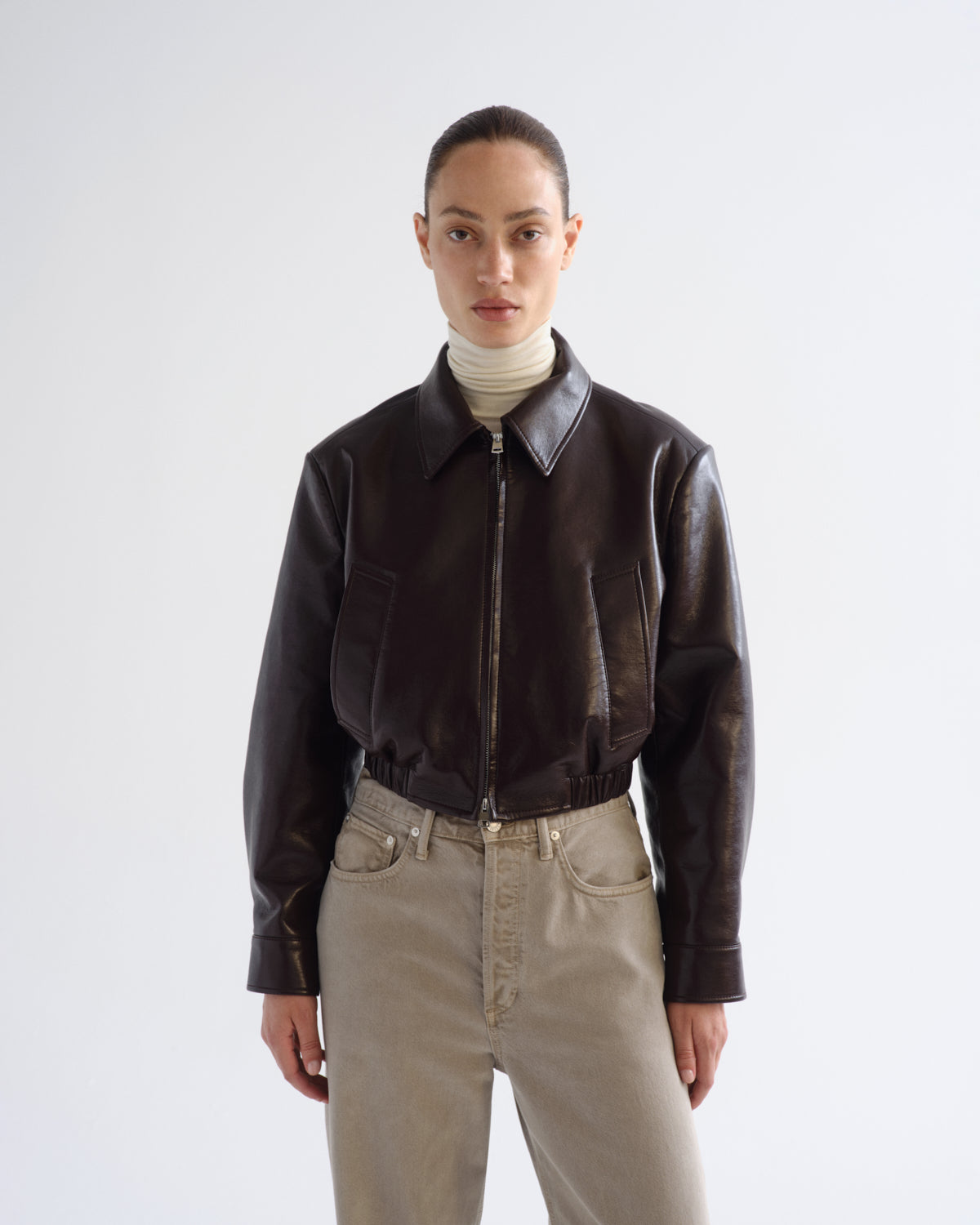 A model wearing a dark brown leather cropped jacket with a front zipper, paired with a cream turtleneck and beige jeans, shot against a light gray studio background.