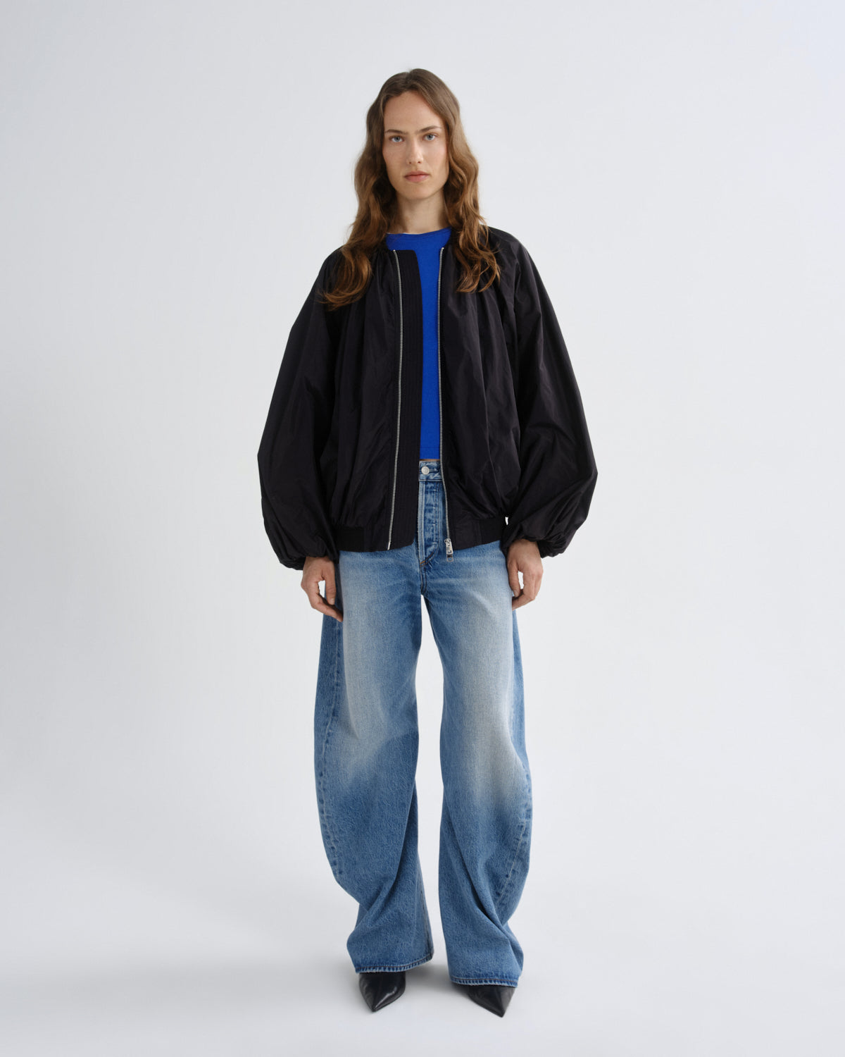 A model stands against a light gray studio background wearing a black bomber jacket over a bright blue top, paired with wide-leg faded blue jeans and black pointed shoes.