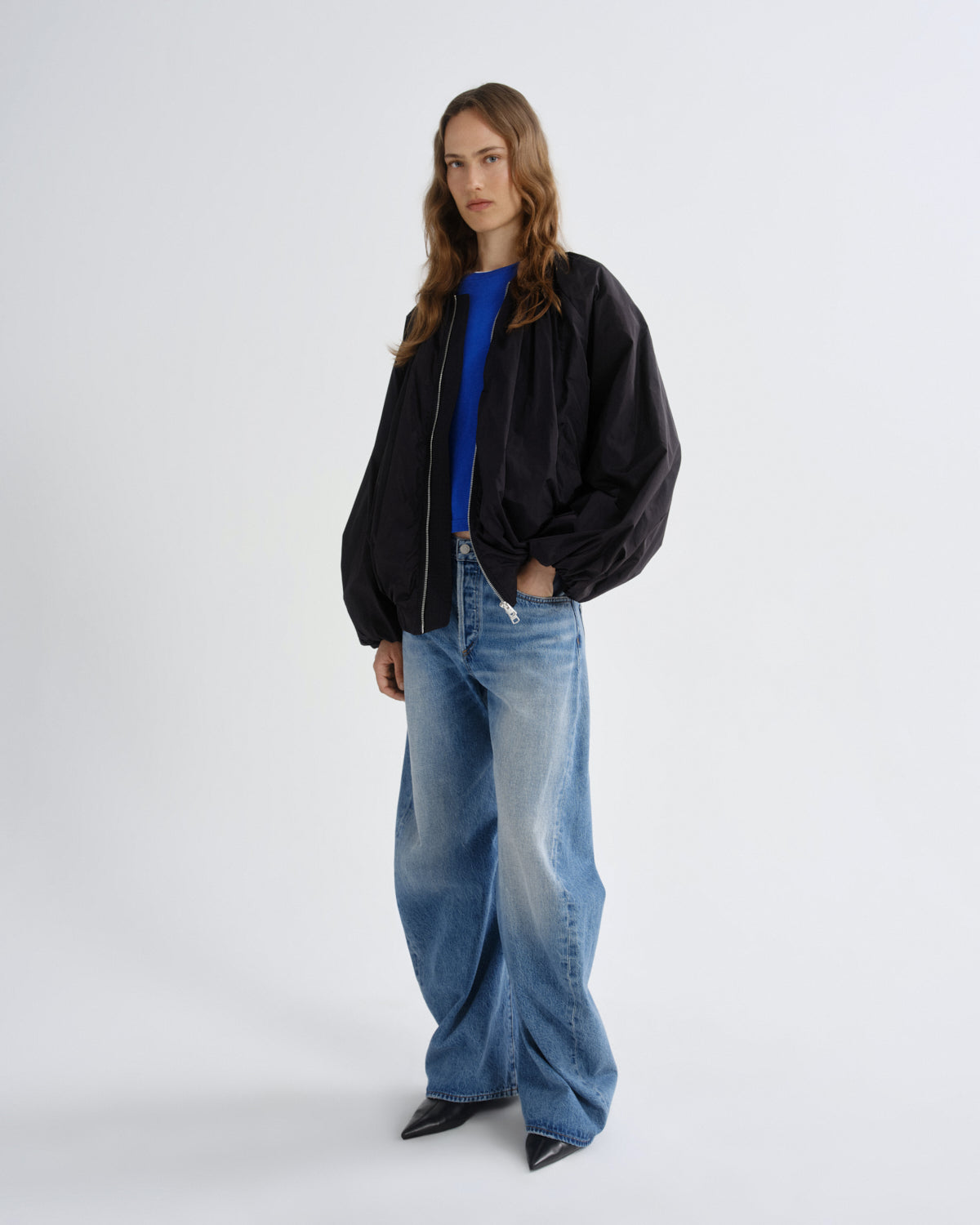 A woman modeling a black bomber jacket over a bright blue top, paired with wide-leg blue jeans and black pointed shoes, against a plain light gray studio background.