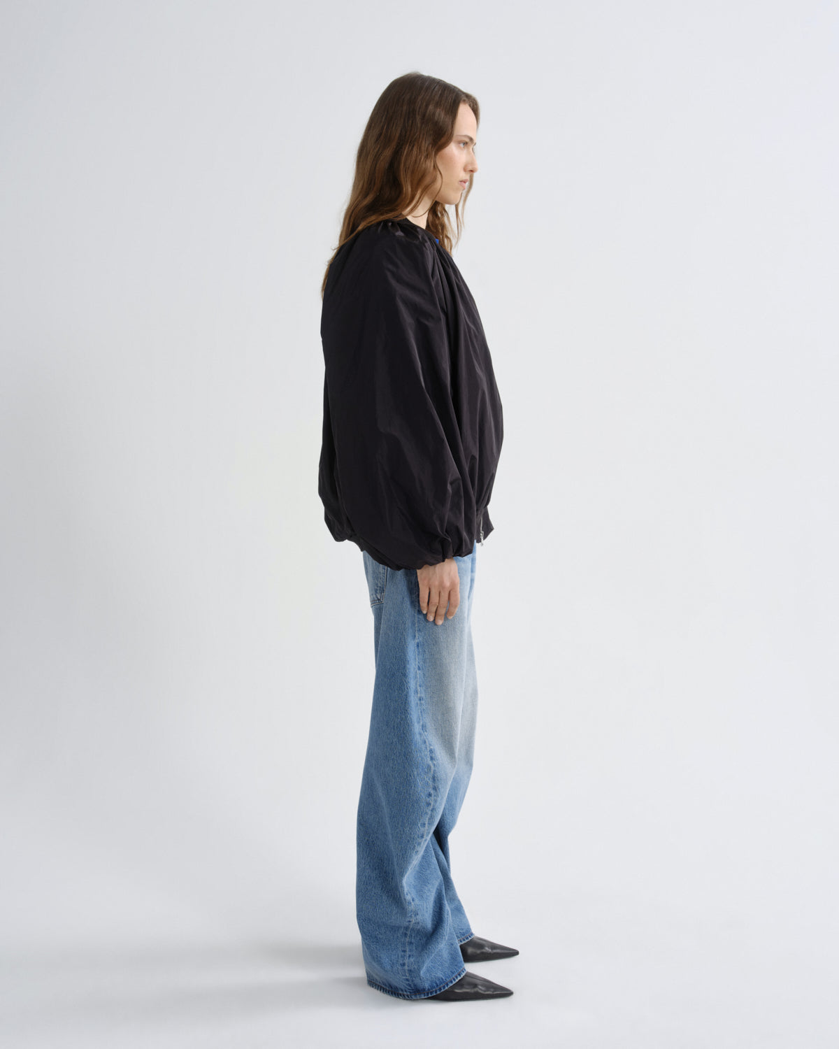A woman stands in profile wearing a black oversized jacket, wide-leg light blue jeans, and black pointed shoes against a plain light gray studio background.