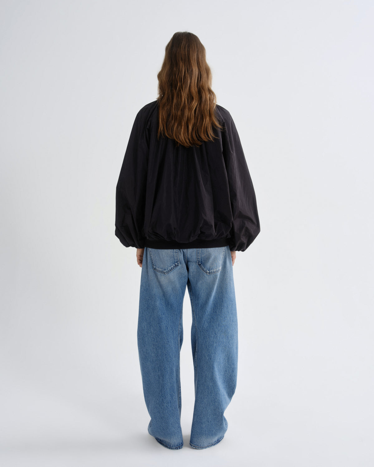 A model viewed from behind wearing a black oversized bomber jacket and wide-leg light blue jeans against a plain light gray studio background.