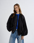 A woman modeling an oversized black bomber jacket with a silver zipper, worn open over a bright blue crewneck shirt and paired with light-wash blue jeans, against a plain light gray studio background.