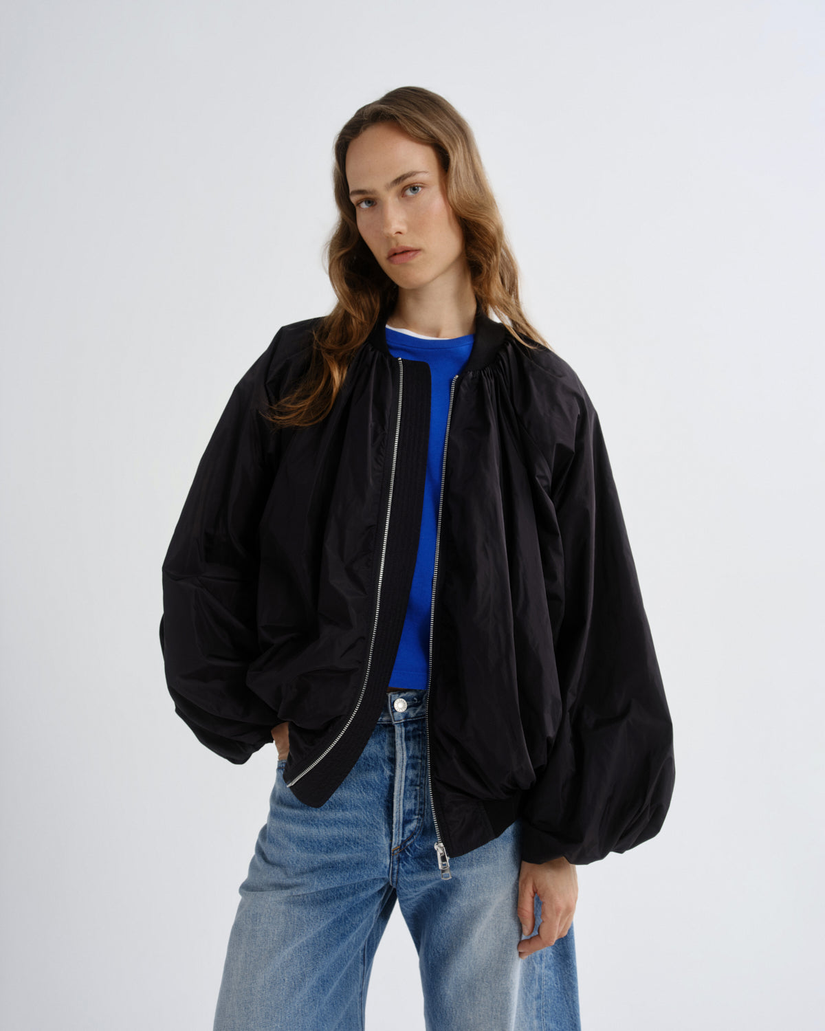 A woman modeling an oversized black bomber jacket with a silver zipper, worn open over a bright blue crewneck shirt and paired with light-wash blue jeans, against a plain light gray studio background.