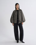 A model stands against a light gray studio background wearing a dark olive green bomber jacket with voluminous balloon sleeves, paired with black wide-leg trousers and black pointed-toe heels.