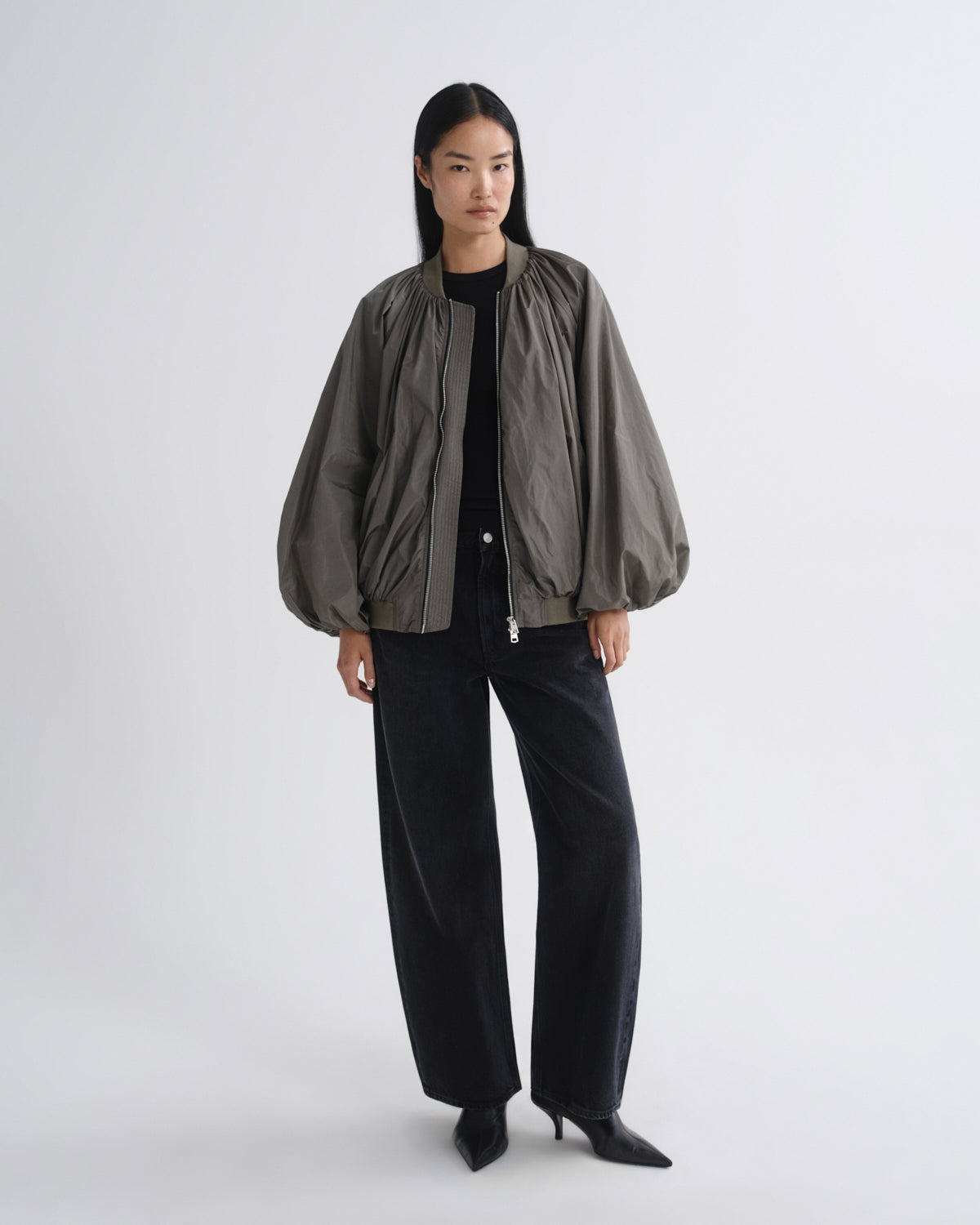 A model stands against a light gray studio background wearing a dark olive green bomber jacket with voluminous balloon sleeves, paired with black wide-leg trousers and black pointed-toe heels.