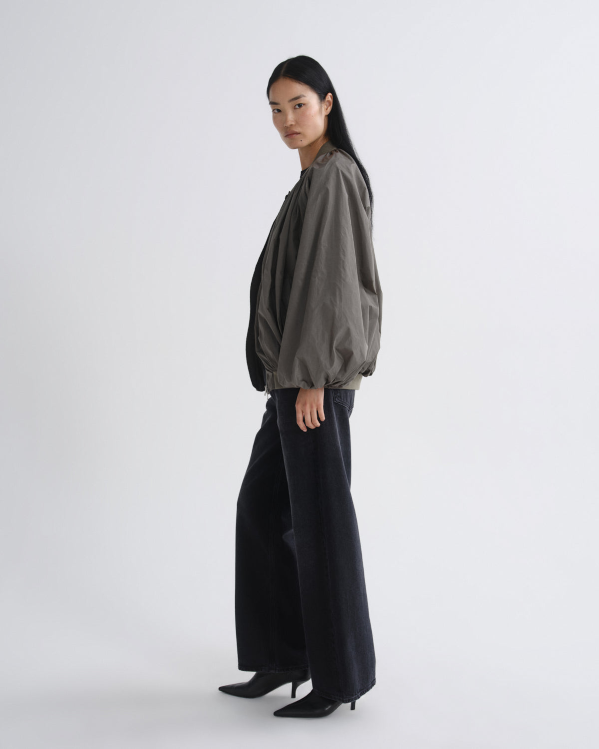 A model is shown in profile wearing a dark olive green balloon-sleeve bomber jacket and wide-leg black trousers against a plain light gray studio background.