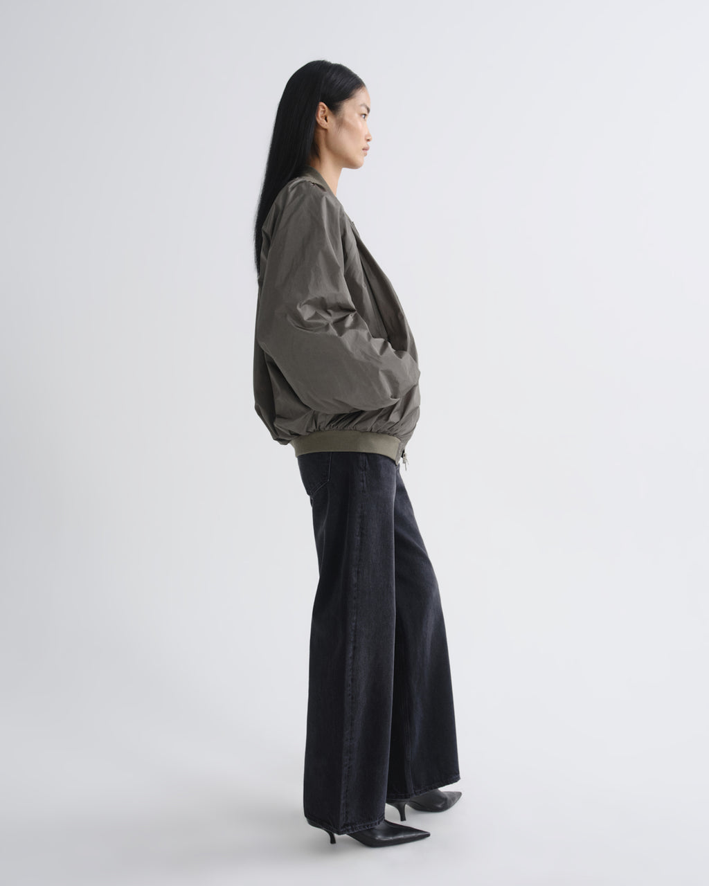 A woman stands in profile wearing an oversized dark olive green jacket with voluminous sleeves and dark wide-leg pants against a plain light gray studio background.