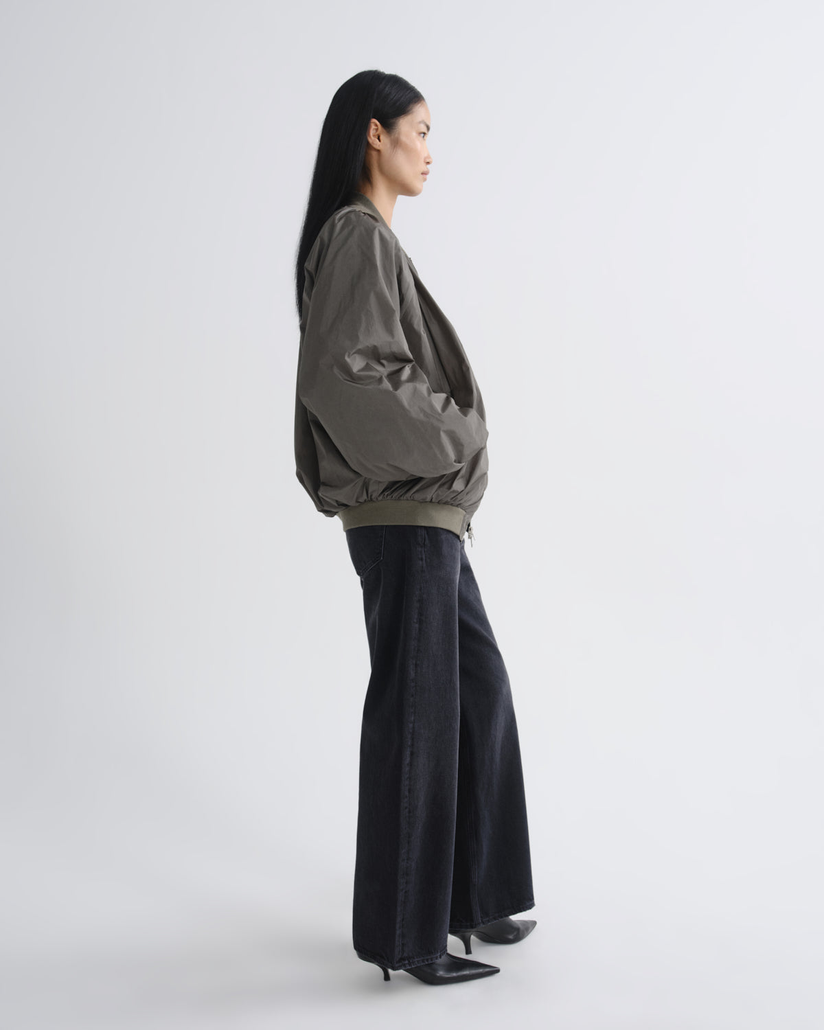 A woman stands in profile wearing an oversized dark olive green jacket with voluminous sleeves and dark wide-leg pants against a plain light gray studio background.