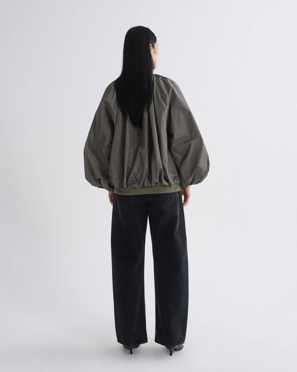 A model shown from the back wearing a dark olive green bomber jacket with voluminous balloon sleeves and black wide-leg jeans, set against a plain light gray studio background.