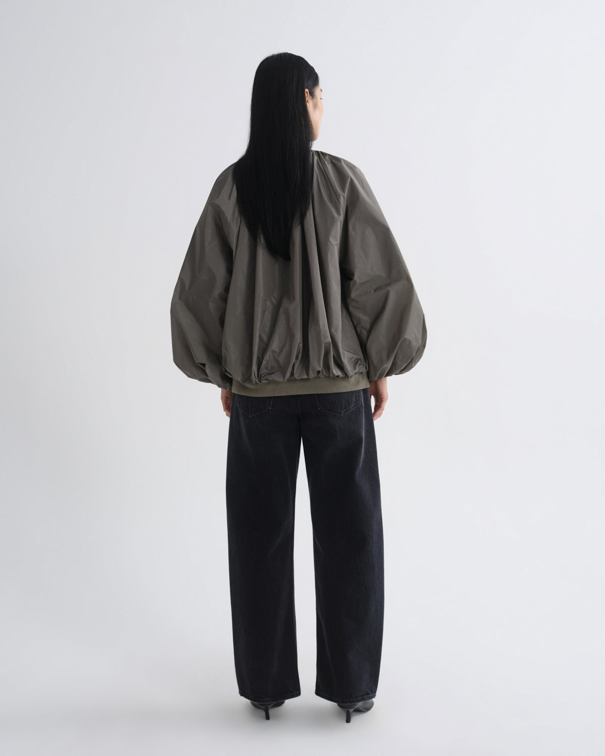 A model shown from the back wearing a dark olive green bomber jacket with voluminous balloon sleeves and black wide-leg jeans, set against a plain light gray studio background.