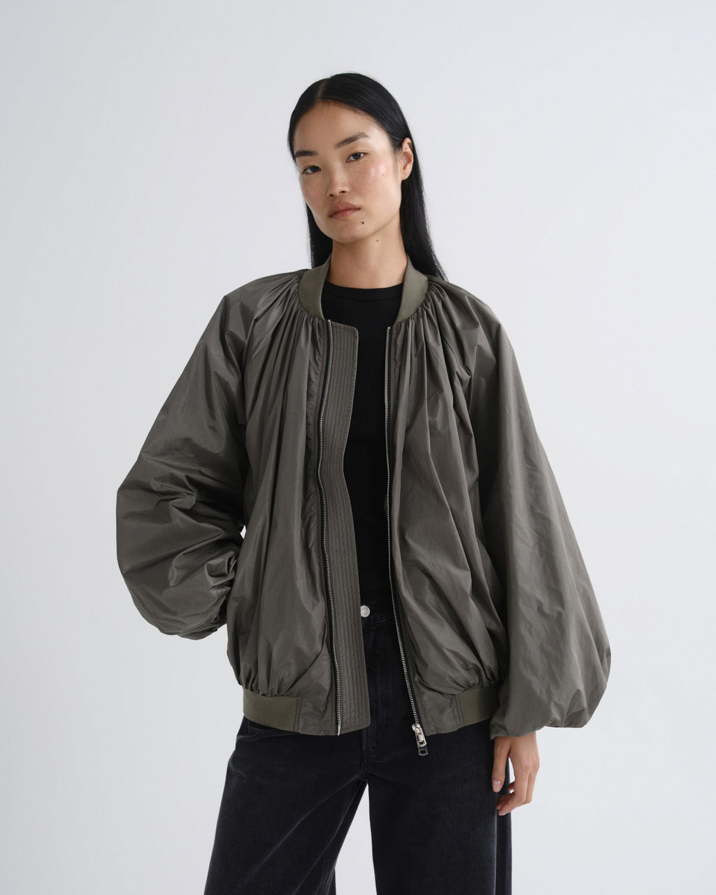 A model wearing a dark olive green bomber jacket with gathered sleeves and a front zipper, styled over a black top and black pants against a light gray studio background.