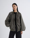A model wearing a dark olive green bomber jacket with gathered sleeves and a front zipper, styled over a black top and black pants against a light gray studio background.