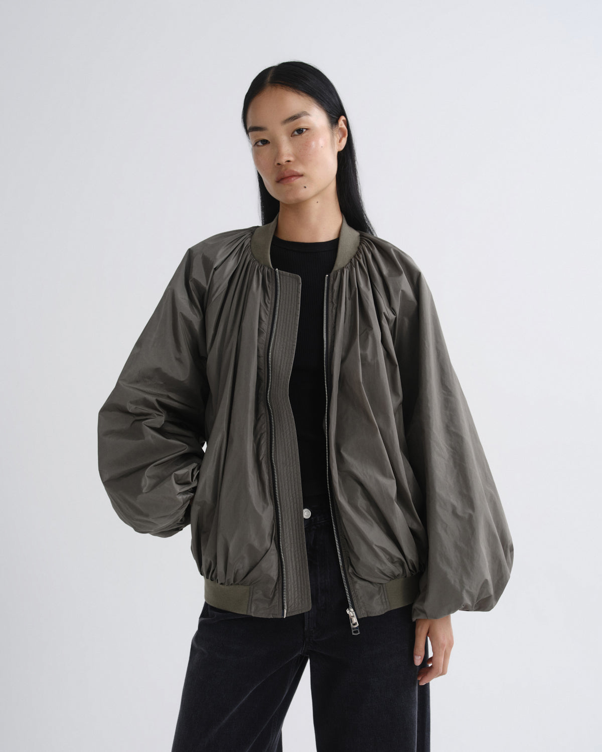 A model wearing a dark olive green bomber jacket with gathered sleeves and a front zipper, styled over a black top and black pants against a light gray studio background.