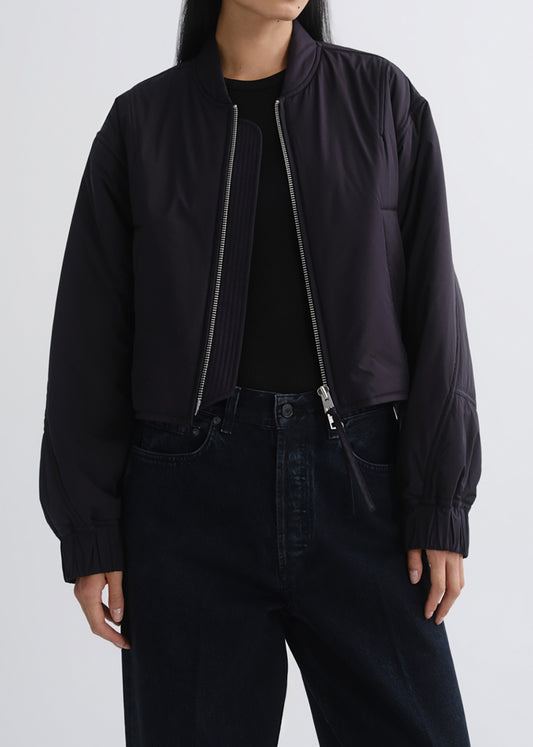 A model wearing a dark navy blue cropped bomber jacket with silver zippers, styled over a black top and dark jeans against a grey background.