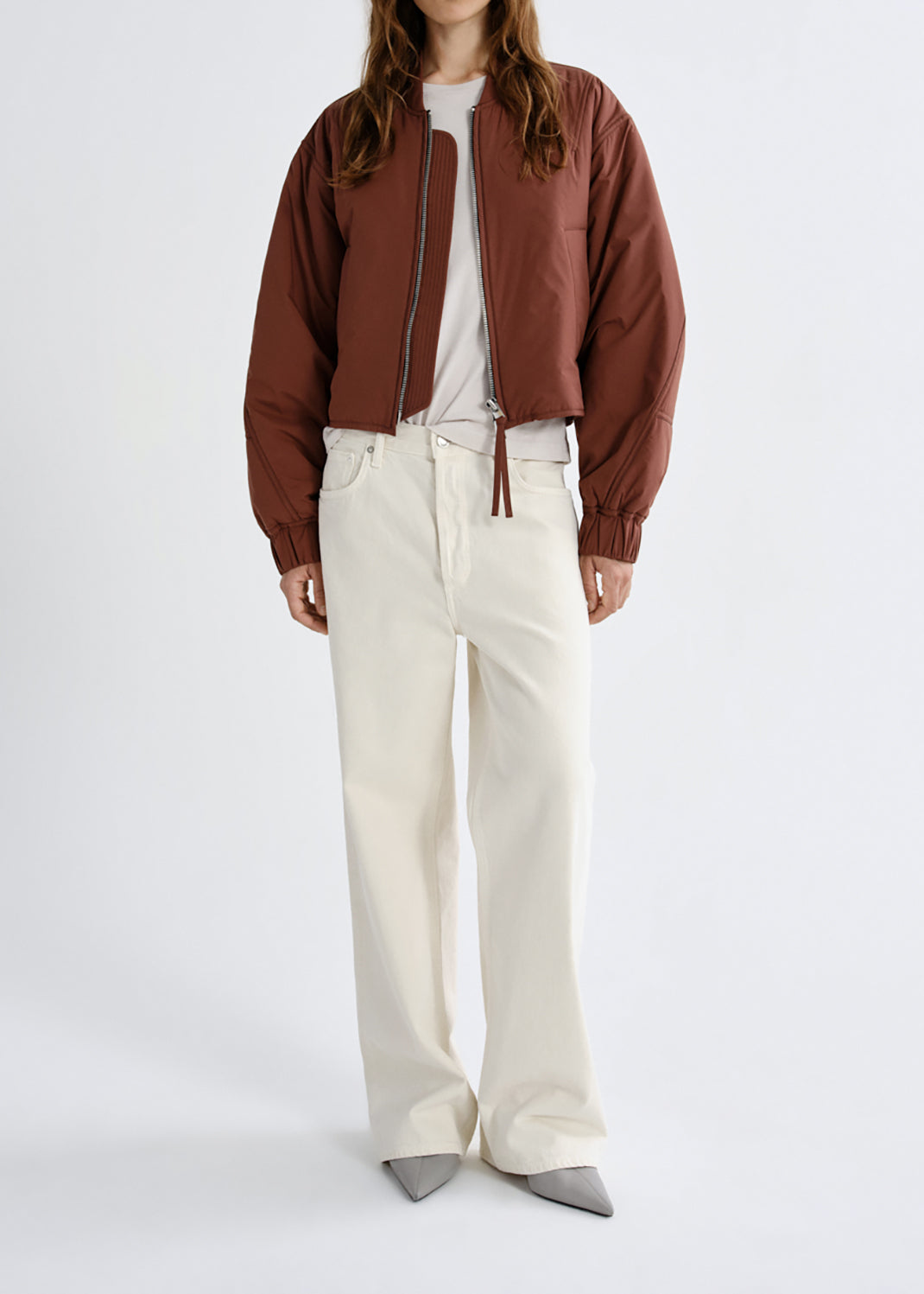 A model wearing a cropped brown bomber jacket, white t-shirt, off-white wide-leg jeans, and grey pointed-toe flats, standing against a grey background.