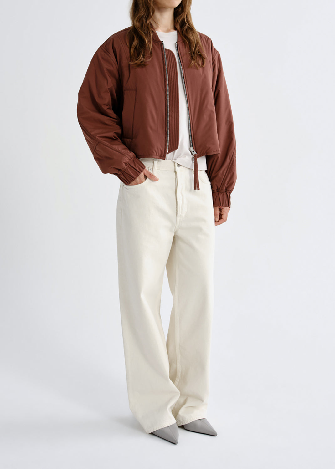A model wearing a brown cropped bomber jacket, off-white wide-leg trousers, a white top, and gray pointed shoes, photographed against a grey background.