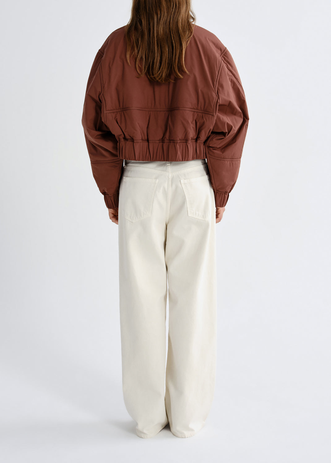 A model shown from the back wearing a cropped brown jacket and cream wide-leg trousers, photographed against a grey background.