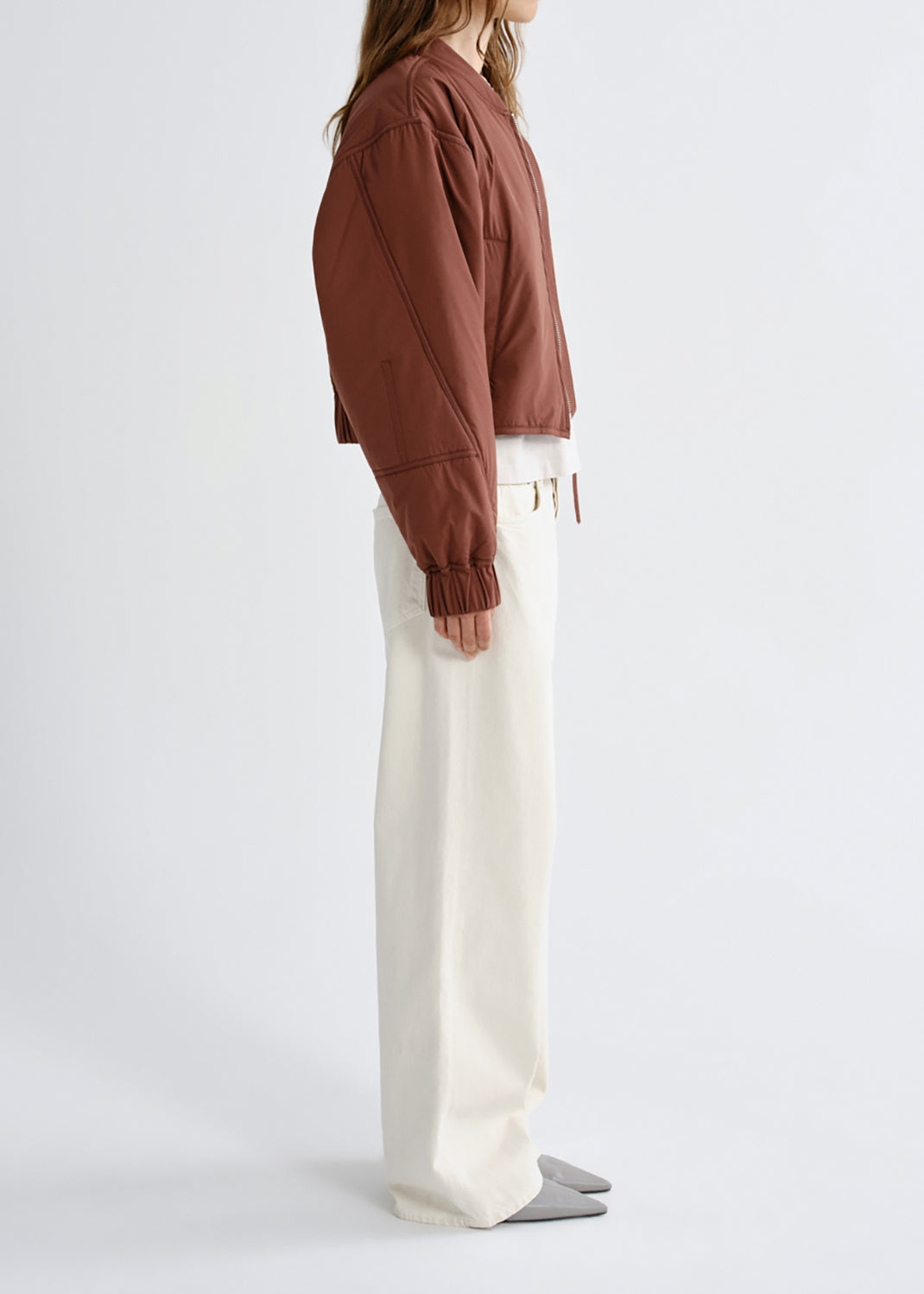 A model shown from the side wearing a cropped brown jacket, off-white wide-leg trousers, and grey slip-on shoes against a grey background.