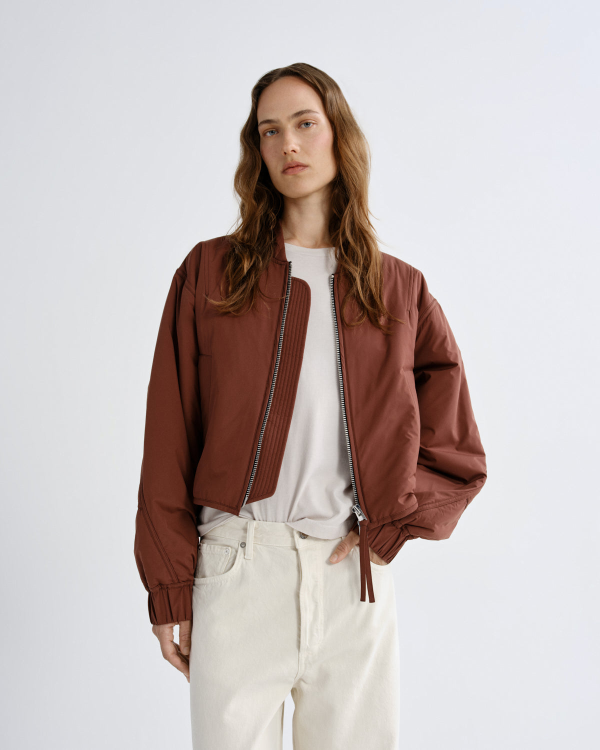 Person wearing a brown jacket over a white shirt and light-colored pants on a plain background
