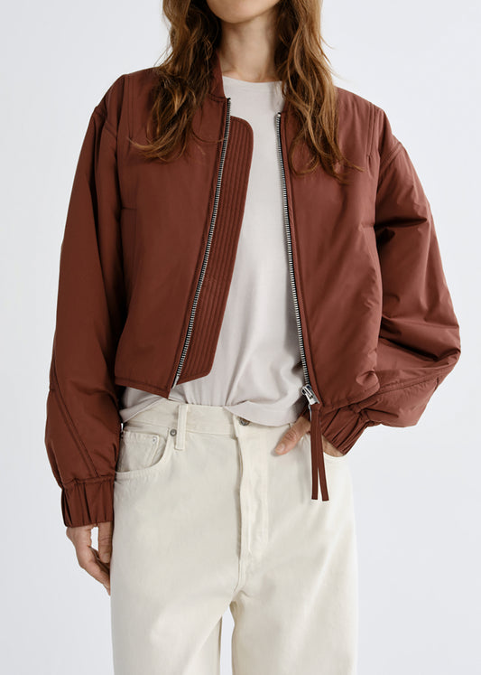 A woman wearing a cropped brown bomber jacket with a silver zipper over a white top and cream-colored pants, shown from the neck down against a grey background.