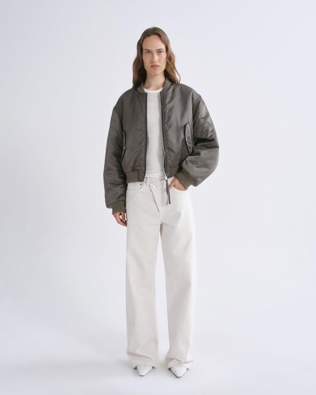 A model stands against a grey background wearing a olive green bomber jacket over a white top and off-white wide-leg trousers.