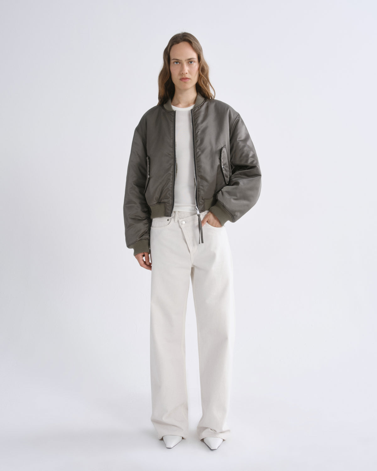 A model stands against a grey background wearing a olive green bomber jacket over a white top and off-white wide-leg trousers.