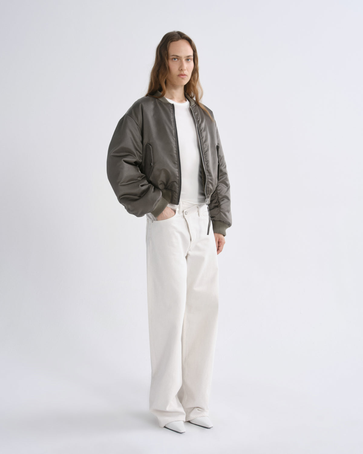 A woman modeling a olive green bomber jacket over a white top and white wide-leg trousers, standing against a grey background.