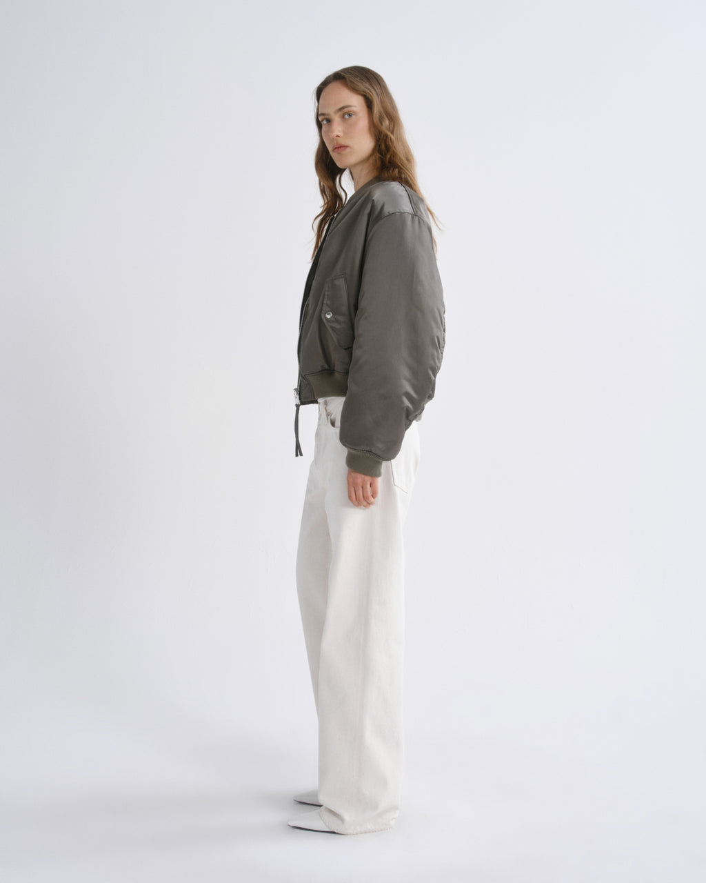 A woman stands in profile wearing a olive green bomber jacket with white wide-leg pants, and white sneakers against a grey background.