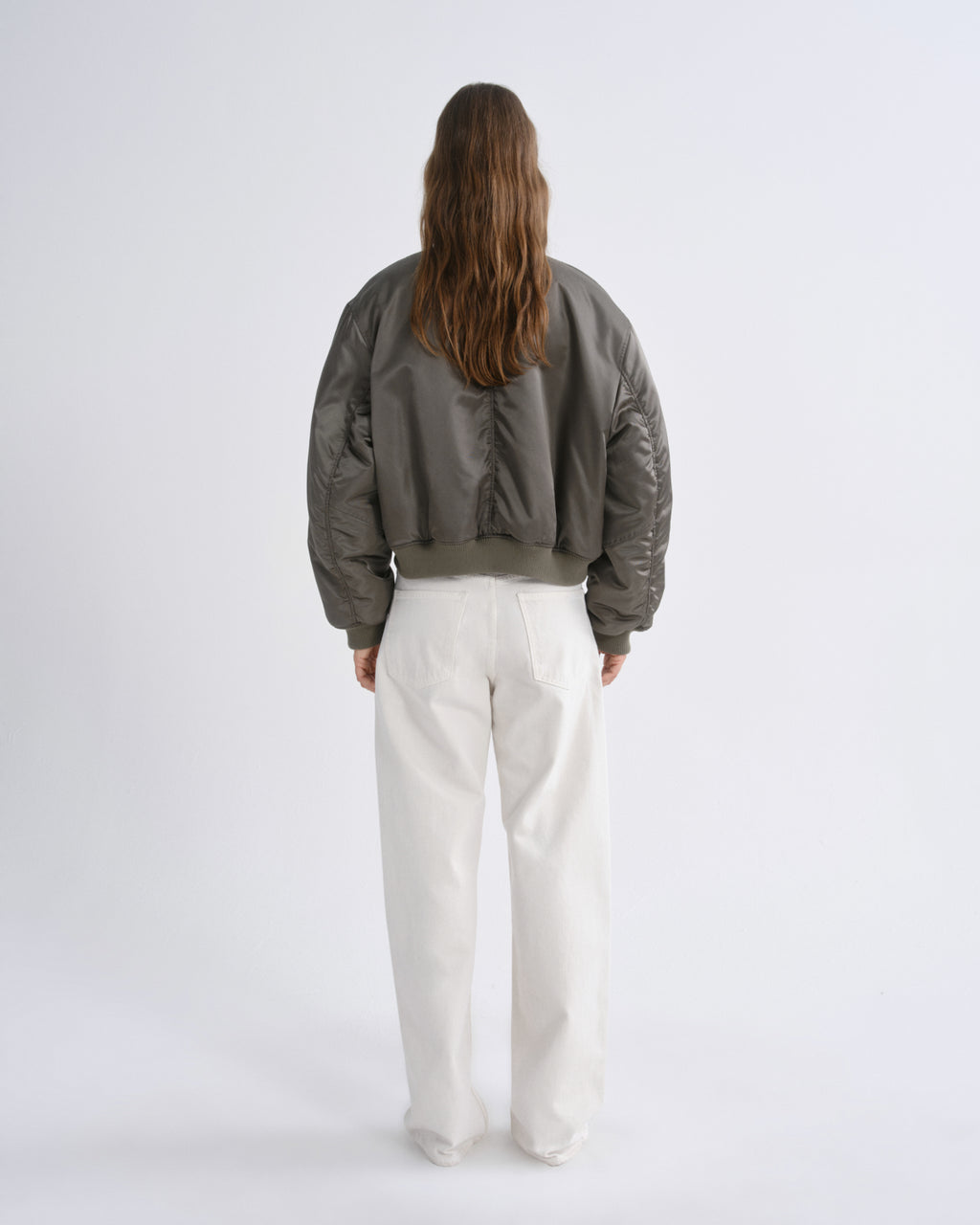 A model viewed from behind wearing a cropped, quilted bomber jacket in a dark olive green color, paired with white wide-leg trousers, against a grey background.