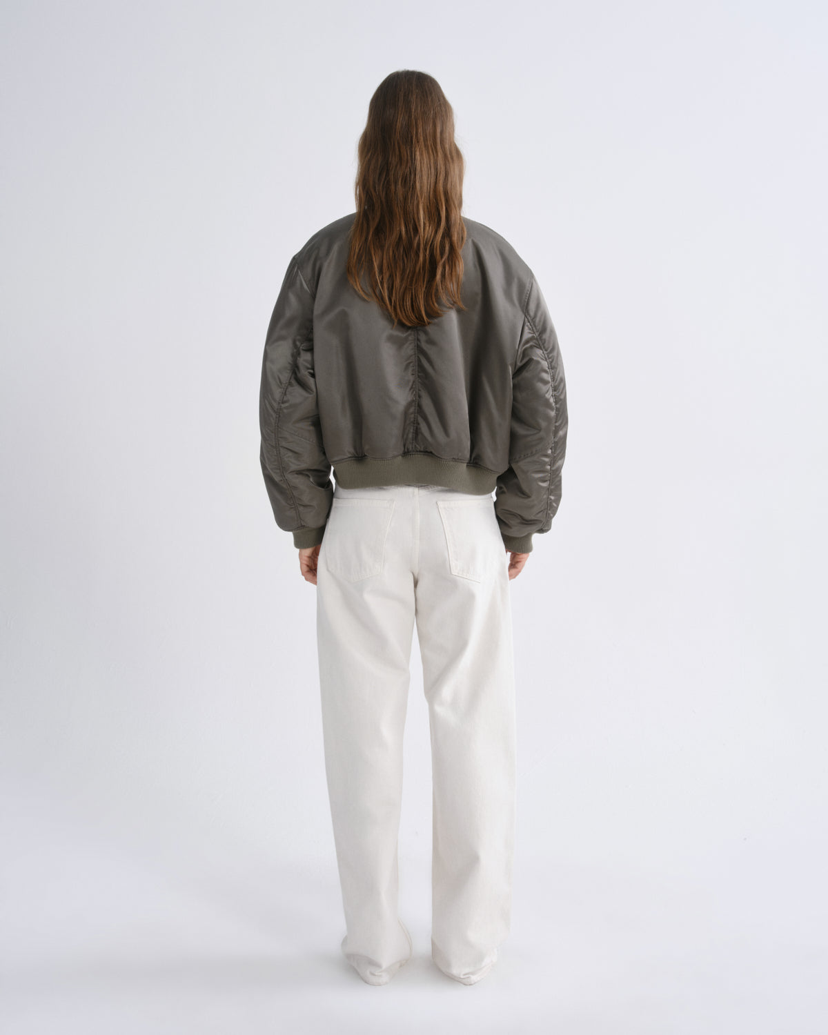 A model viewed from behind wearing a cropped, quilted bomber jacket in a dark olive green color, paired with white wide-leg trousers, against a grey background.